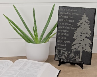 May include: A black slate plaque with a tree design and the text "He is like a tree planted by streams of water...Psalm 1:3". A potted aloe vera plant and an open book are also in the image.