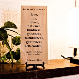 May include: A wooden plaque with the text "But the fruit of the Spirit is love, joy, peace, patience, kindness, goodness, faithfulness, gentleness, self-control;" and "Galatians 5:22-23". The plaque is on a wooden shelf with a lamp and a plant.
