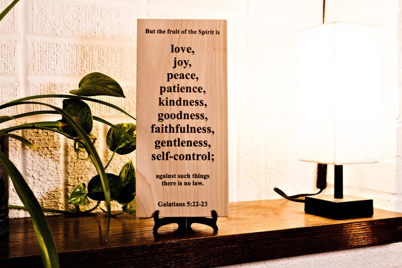 May include: A wooden plaque with the text "But the fruit of the Spirit is love, joy, peace, patience, kindness, goodness, faithfulness, gentleness, self-control;" and "Galatians 5:22-23". The plaque is on a wooden shelf with a lamp and a plant.