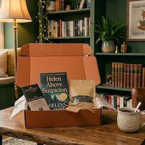 May include: An open brown cardboard box filled with a book titled "Helen Above Suspicion", a chocolate bar, and a bag of tea. A mug with a leaf design sits on a wooden table. The background includes a bookshelf and a window.