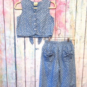 May include: A blue denim vest and pants set with a white floral pattern. The vest has a button closure and ties at the waist. The pants have a relaxed fit and pockets.