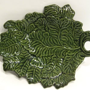 May include: A green ceramic serving tray shaped like a leaf with a handle. The tray has a textured surface that resembles a leaf.