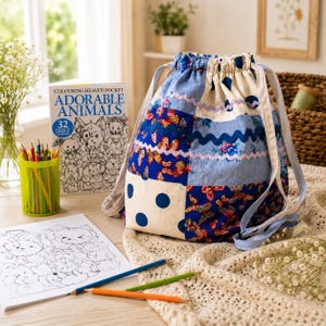 May include: A patchwork drawstring bag with blue, white, and floral patterns. The bag is next to a colouring book titled "Colouring Heaven Pocket Adorable Animals" and a cup of coloured pencils. A colouring page with animal illustrations and coloured pencils are on the table.