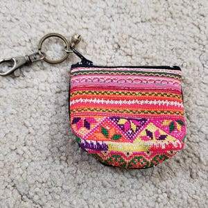 May include: A small, colorful coin purse with a zipper closure and a metal keychain. The purse features a vibrant, patterned design with pink, orange, yellow, and purple hues. The keychain includes a clasp and a small bell.