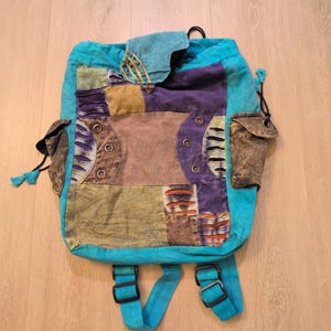 May include: A patchwork backpack in shades of teal, purple, brown, and olive green. The bag features multiple pockets, adjustable straps, and a drawstring closure. The design incorporates various fabric textures and patterns, creating a bohemian aesthetic.