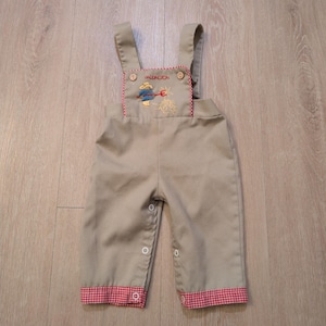 May include: Tan baby overalls with red gingham trim and straps. The bib features an embroidered design with the text "Paddington". The overalls have snap closures on the sides and at the bottom of the legs.
