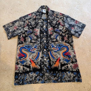 May include: Short-sleeved button-down shirt with a black background and a dragon design. The dragon is blue, orange, and red. The shirt has a floral pattern in gray, red, and black.