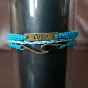 May include: Three handmade bracelets. The top bracelet is turquoise blue with a gold-colored metal plate that reads "Love". The middle bracelet is white and turquoise blue. The bottom bracelet has a gold-colored metal clasp.