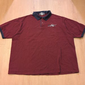 May include: A maroon polo shirt with a navy blue collar and sleeve trim. The shirt features a small embroidered design on the chest. The shirt is made of a soft, breathable material.