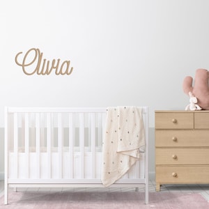 Peut inclure: Un lit b&eacute;b&eacute; blanc avec une couverture blanche et une plaque de nom en bois qui dit "Olivia" au-dessus. Une commode en bois avec un cactus rose et un lapin blanc sur le dessus est &agrave; droite du lit b&eacute;b&eacute;.