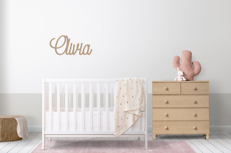 Peut inclure: Un lit b&eacute;b&eacute; blanc avec une couverture blanche et une plaque de nom en bois qui dit "Olivia" au-dessus. Une commode en bois avec un cactus rose et un lapin blanc sur le dessus est &agrave; droite du lit b&eacute;b&eacute;.
