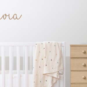 Peut inclure: Un lit b&eacute;b&eacute; blanc avec une couverture cr&egrave;me drap&eacute;e sur le c&ocirc;t&eacute;. Le nom "Olivia" est &eacute;crit sur le mur au-dessus du lit b&eacute;b&eacute; en &eacute;criture brune. Une commode en bois avec cinq tiroirs se trouve &agrave; droite de l'image. Un cactus rose et un lapin blanc sont pos&eacute;s sur la commode.