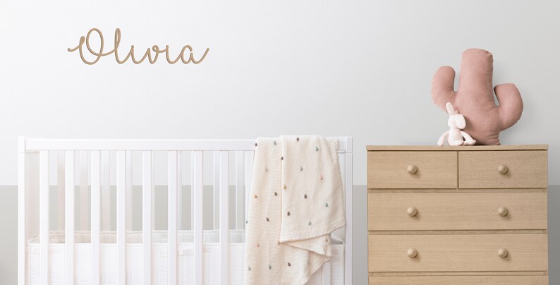 Peut inclure: Un lit b&eacute;b&eacute; blanc avec une couverture cr&egrave;me drap&eacute;e sur le c&ocirc;t&eacute;. Le nom "Olivia" est &eacute;crit sur le mur au-dessus du lit b&eacute;b&eacute; en &eacute;criture brune. Une commode en bois avec cinq tiroirs se trouve &agrave; droite de l'image. Un cactus rose et un lapin blanc sont pos&eacute;s sur la commode.