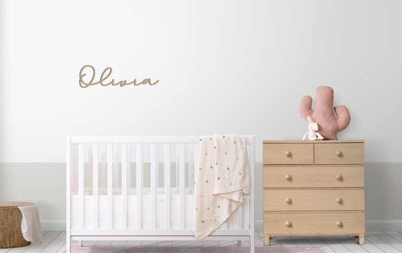 Peut inclure: Un lit b&eacute;b&eacute; blanc avec une couverture blanche et une commode en bois surmont&eacute;e d'un cactus rose et d'un lapin blanc. Le mur est blanc avec le nom "Olivia" &eacute;crit en script brun.