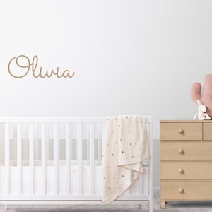 Peut inclure: Un lit b&eacute;b&eacute; blanc avec une couverture blanche et une commode en bois surmont&eacute;e d'un cactus rose et d'un lapin en peluche. Le mur est blanc avec le nom "Olivia" &eacute;crit en cursive brune.
