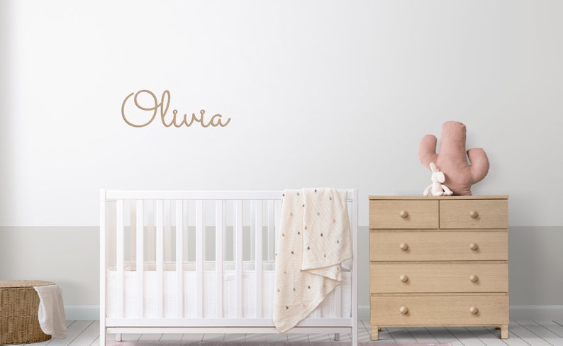 Peut inclure: Un lit b&eacute;b&eacute; blanc avec une couverture blanche et une commode en bois surmont&eacute;e d'un cactus rose et d'un lapin en peluche. Le mur est blanc avec le nom "Olivia" &eacute;crit en cursive brune.