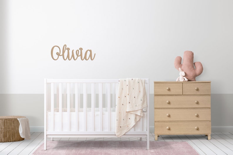 Peut inclure: Un lit b&eacute;b&eacute; blanc avec une couverture blanche et une commode en bois &agrave; six tiroirs. Un coussin en forme de cactus rose est pos&eacute; sur la commode. Le nom "Olivia" est &eacute;crit sur le mur en cursive.
