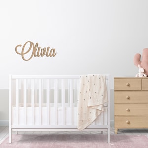 Peut inclure: Un lit b&eacute;b&eacute; blanc avec une couverture beige et une commode en bois surmont&eacute;e d'un jouet cactus rose. Le mur au-dessus du lit b&eacute;b&eacute; porte une plaque de nom en bois sur laquelle est inscrit "Olivia".