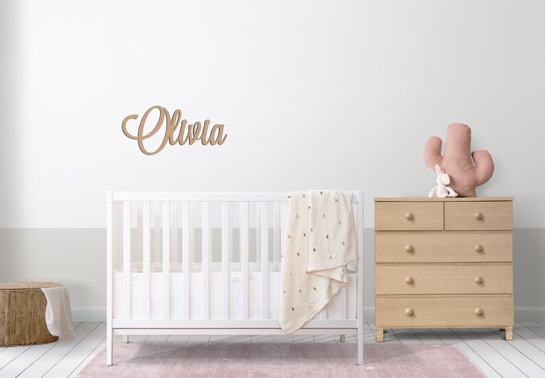 Peut inclure: Un lit b&eacute;b&eacute; blanc avec une couverture beige et une commode en bois surmont&eacute;e d'un jouet cactus rose. Le mur au-dessus du lit b&eacute;b&eacute; porte une plaque de nom en bois sur laquelle est inscrit "Olivia".