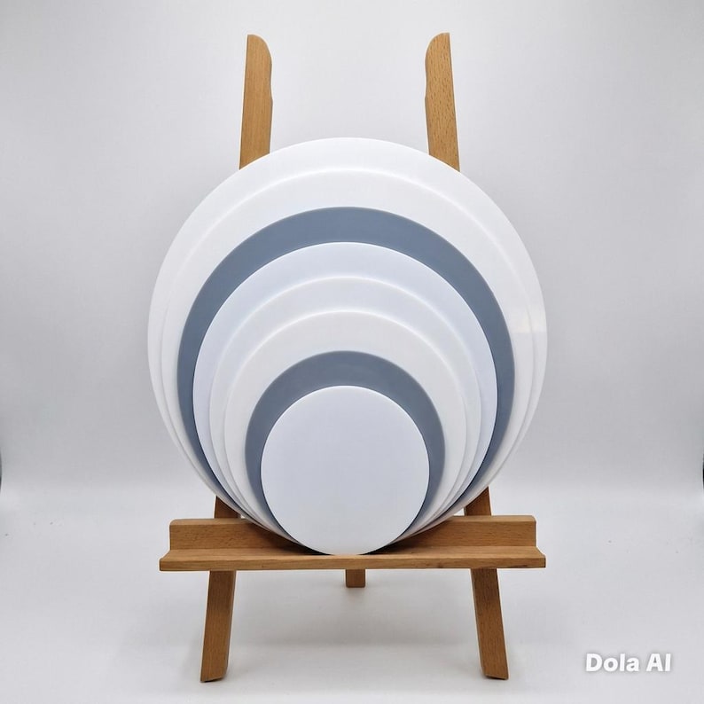 K&ouml;nnte beinhalten: Ein geschichtetes, kreisf&ouml;rmiges Kunstwerk mit abwechselnden wei&szlig;en und grauen B&auml;ndern, pr&auml;sentiert auf einer kleinen Holzstaffelei. Das Design zeigt konzentrische Kreise, die einen dreidimensionalen Effekt erzeugen. Das Kunstwerk ist vor einem wei&szlig;en Hintergrund platziert.