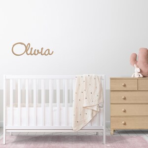 Peut inclure: Un lit b&eacute;b&eacute; blanc avec une couverture blanche et une pancarte en bois avec le nom "Olivia" au-dessus. Une commode en bois avec cinq tiroirs est &agrave; droite du lit b&eacute;b&eacute;. Un jouet cactus rose est pos&eacute; sur la commode.