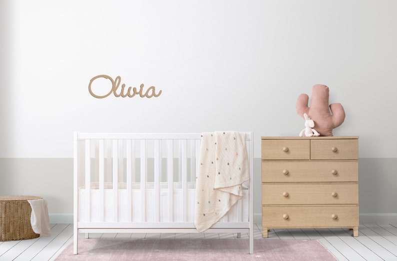 Peut inclure: Un lit b&eacute;b&eacute; blanc avec une couverture blanche et une pancarte en bois avec le nom "Olivia" au-dessus. Une commode en bois avec cinq tiroirs est &agrave; droite du lit b&eacute;b&eacute;. Un jouet cactus rose est pos&eacute; sur la commode.