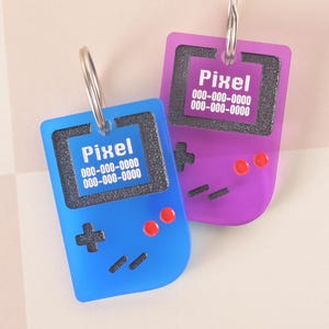 Puede incluir: Dos llaveros acrílicos con forma de consolas de juegos portátiles retro. Uno es azul, el otro morado. Cada uno tiene un llavero plateado y la palabra "Pixel" impresa en la pantalla, junto con un número de teléfono. Los llaveros tienen detalles negros y botones rojos.