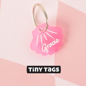 May include: Pink seashell-shaped pet tag with a silver ring and the name "Gracie" written in white. The tag is on a pink and white checkered background with the words "TINY TAGS" printed in black.