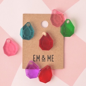 May include: A selection of colourful, faceted gemstone-shaped charms in shades of pink, teal, green, purple, and red. The charms are displayed on a brown card with the text "EM & ME". The background features a pink and white geometric pattern.