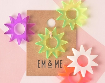 May include: Five colorful star-shaped rings with a hole in the center. The rings are made of translucent plastic and are in shades of purple, green, white, and orange. The rings are arranged on a brown paper tag with the text "EM & ME" printed on it.