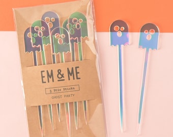 May include: A package of six iridescent ghost-shaped stir sticks. The package is brown with the text "EM & ME 6 Stir Sticks GHOST PARTY emandmestudio.com". Two individual ghost stir sticks are shown outside of the package.