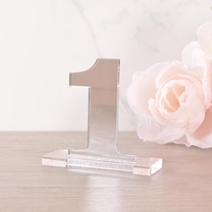 Può includere: Un numero di tavolo in acrilico argentato numero uno con una base in acrilico trasparente. Il numero è su una superficie in legno chiaro.