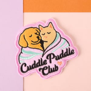 May include: Embroidered patch featuring a golden retriever and a cat snuggled together under a pink blanket with a mint green trim. The patch reads "Cuddle Puddle Club" in a black script font with a small orange heart.