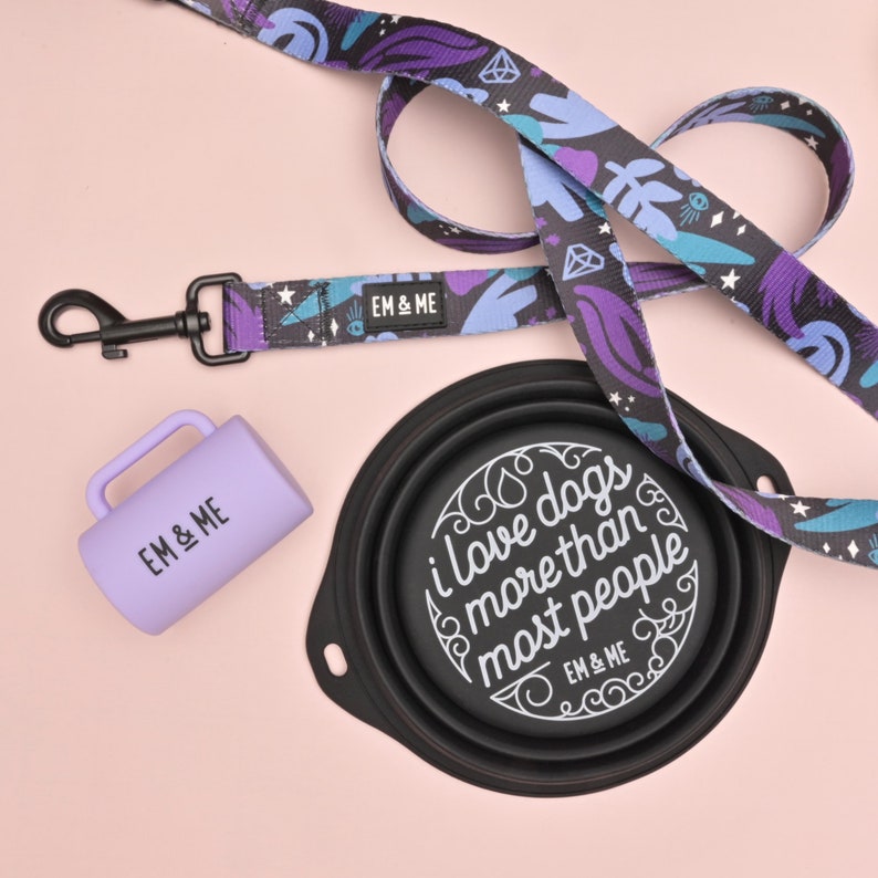 May include: A purple and blue patterned dog leash with a matching purple water bowl and a purple travel mug. The water bowl is collapsible and has the text "I love dogs more than most people" printed on it. All items have the text "EM & ME" printed on them.
