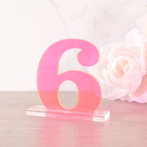 Può includere: Numero di tavolo sei in acrilico rosa su un supporto in acrilico trasparente. Il numero è contornato in giallo.