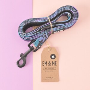 May include: A purple and blue patterned dog leash with a black metal clip and a brown tag that says "EM & ME Pet Accessory SPOOKY MAGIC CHICAGO, IL em&mestudio.com" and "M".