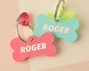 Puede incluir: Dos etiquetas para perros en forma de hueso, una rosa y otra azul, con el nombre "Roger" impreso en cada una. Las etiquetas están unidas a llaveros plateados.