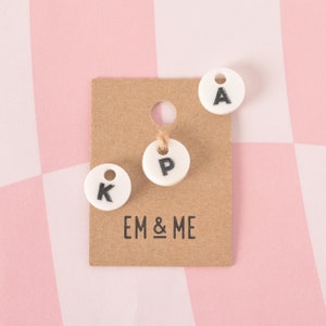 Op de afbeelding: Drie witte ronde labels met zwarte letters op een bruine kaart. De letters zijn K, P en A. De kaart heeft de tekst "EM & ME" erop gedrukt.