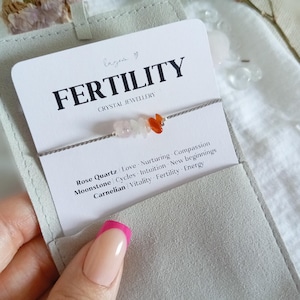 May include: A delicate bracelet featuring small, tumbled rose quartz, moonstone, and carnelian crystal chips on a thin grey cord. The bracelet is attached to a white display card labeled FERTILITY CRYSTAL JEWELLERY with text listing the stones.