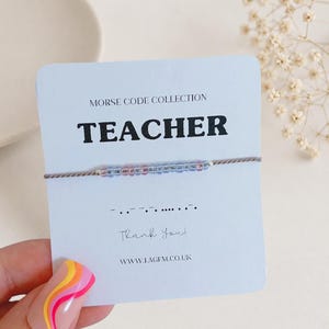 May include: A Morse code bracelet featuring small blue and pink beads on a thin grey cord, displayed on a white card labelled MORSE CODE COLLECTION TEACHER with the corresponding Morse code dots and dashes and the text Thank You and WWW.LAGEM.CO.UK.