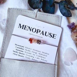 May include: A delicate bracelet featuring small, multi-colored crystal chips on a thin grey cord, displayed on a white card labeled MENOPAUSE CRYSTAL JEWELLERY. The card lists Moonstone, Carnelian, Sunstone, Smoky Quartz, and Clear Quartz with their associated properties, all tucked into a light grey fabric pouch.