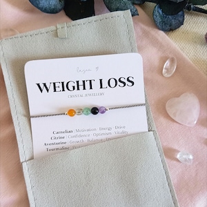 Puede incluir: Una pulsera de hilo gris fino con cinco peque&ntilde;as cuentas redondas de piedras preciosas en naranja, transparente, verde, negro y morado, presentada en una tarjeta blanca con el texto WEIGHT LOSS CRYSTAL JEWELLERY y una lista de propiedades.
