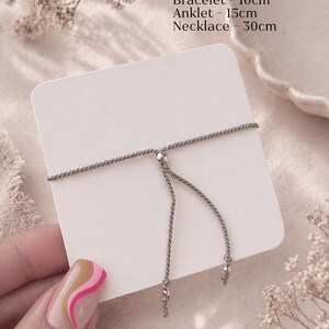 Puede incluir: Una pulsera de cord&oacute;n gris con cierre de cuenta met&aacute;lica deslizante sobre una tarjeta blanca. El texto dice: Jewellery length: Bracelet - 10cm, Anklet - 15cm, Necklace - 30cm. Abajo, el texto dice: Adjustable universal size.