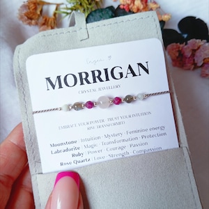 May include: A delicate beaded bracelet featuring a central rose quartz bead flanked by ruby and labradorite stones on a thin cord. The bracelet is displayed on a white card labeled MORRIGAN CRYSTAL JEWELLERY with text listing moonstone, labradorite, ruby, and rose quartz properties.