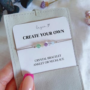 May include: A thin silver-toned chain bracelet featuring five small, multi-coloured round crystal beads in green, yellow, purple, and light blue. The jewellery is displayed on a white card with the text "CREATE YOUR OWN CRYSTAL BRACELET ANKLET OR NECKLACE" inside a light grey fabric pouch.