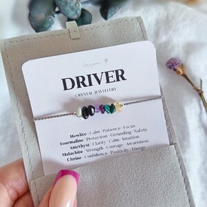 May include: A delicate bracelet featuring small, polished gemstone chips in white, black, purple, green, and yellow, strung on a thin grey cord. The bracelet is displayed on a white card labeled DRIVER CRYSTAL JEWELLERY with a list of stone names.