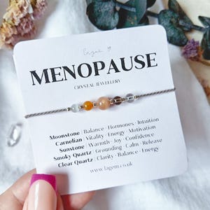 May include: A delicate bracelet on a white display card labeled MENOPAUSE CRYSTAL JEWELLERY. The bracelet features five small beads: moonstone, carnelian, sunstone, smoky quartz, and clear quartz, strung on a thin grey cord with metallic spacers.