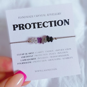 May include: A crystal bracelet featuring clear quartz, amethyst, labradorite, and tourmaline beads. The card displays "PROTECTION" and crystal properties. The bracelet is strung on a thin, neutral-toned cord.