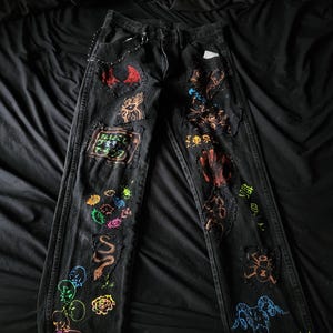 May include: Black denim jeans with colorful hand-painted designs, including a snake, a skull, and Japanese characters. The jeans have several patches and rips.