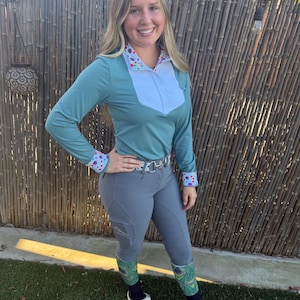 May include: A long-sleeved teal shirt with a white chest panel and a colorful floral print on the collar and cuffs. The shirt is paired with grey riding breeches featuring a side pocket, a silver-buckled belt, and green peacock-patterned socks.